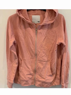 Rosa hoodie med dragkedja från 157 - Säljer en ljusrosa hoodie från 157 med dragkedja framtill och två stora fickor. Hoodien har huva och är tillverkad i mjukt bomullsmaterial. Perfekt för en avslappnad och bekväm stil.