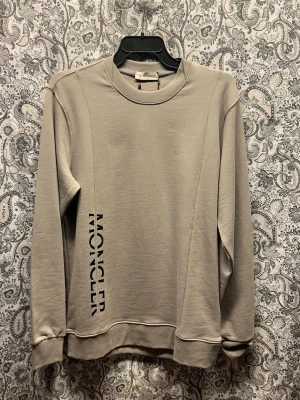 Grå sweatshirt från Moncler - Snygg grå sweatshirt från Moncler med svart logotyp tryckt vertikalt på vänster sida. Tröjan har rund halsringning, långa ärmar och ribbade muddar vid ärmslut och nederkant. Tillverkad i mjukt bomullsmaterial för en bekväm känsla.