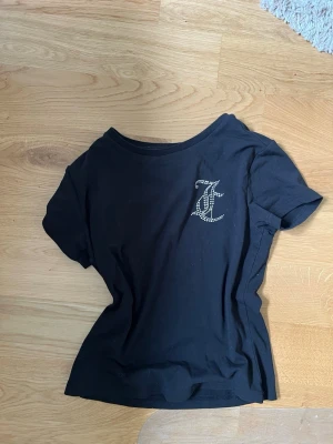 Svart t-shirt med strassdetalj - Snygg svart t-shirt med korta ärmar och en glittrig strassbokstav på bröstet. T-shirten har en klassisk rund halsringning och är gjord i mjukt bomullsmaterial. Aldrig använd men för liten så därför jag säljer.