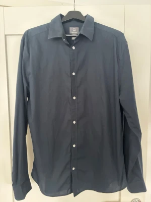Mörkblå skjorta från H&M - Klassisk mörkblå skjorta från H&M med lång ärm och vita knappar framtill. Skjortan har en enkel, stilren design och är tillverkad i mjuk bomull som känns skön mot huden. Perfekt för dig som gillar en clean och tidlös look.