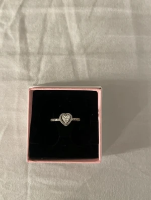 Silverring med hjärta från Pandora - Säljer en snygg silverring från Pandora med ett hjärta i mitten, dekorerad med gnistrande stenar runt hjärtat och längs ringen. Kommer i original ask. Perfekt för dig som gillar stilrena och romantiska smycken.