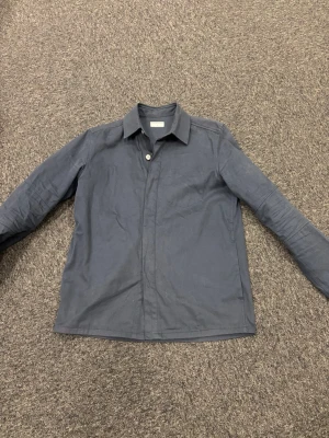 Mörkblå overshirt från Tiger of Sweden - Stilren mörkblå overshirt från Tiger of Sweden i bomullstyg. Klassisk krage, dolda knappar framtill och långa ärmar. Perfekt lager på lager-plagg för en clean och modern look.