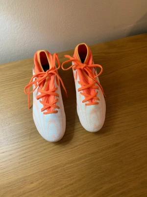 Nike vita och orange fotbollsskor - Säljer ett par snygga fotbollsskor från Nike i vitt och orange. Skorna har snörning, låg profil och mönstrad ovandel för bra bollkontroll. Utrustade med fasta dobbar för spel på naturgräs. Perfekta för dig som vill sticka ut på planen!