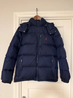 Mörkblå pufferjacka från Polo Ralph Lauren - Säljer en mörkblå pufferjacka från Polo Ralph Lauren i storlek XL. Jackan har hög krage, avtagbar huva och flera dragkedjeförsedda fickor. Klassisk röd logga på bröstet och elastiska muddar vid ärmsluten. Perfekt för kalla dagar. Passar inte på mig och jag är M då rekommenderar till dom som har S