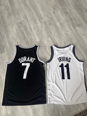 Brooklyn Nets basket jersey Durant & Irving - Två basketlinnen från Brooklyn Nets, ett svart med vitt tryck 'Durant 7' och ett vitt med svart tryck 'Irving 11'. Klassisk ärmlös modell med rund hals och kontrastfärgade kanter. Tillverkade i lätt och ventilerande polyester, perfekta för basket eller streetwear. Vita i storlek S och svarta i storlek M. 250kr per jersey, 400kr för båda!