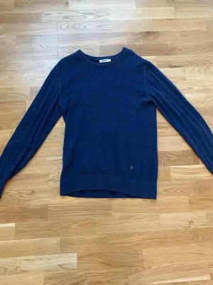 Stickad tröja J.Lindeberg - Mörkblå stickad tröja från Gant med diskret logga nedtill. Tröjan har rund halsringning, långa ärmar och ett klassiskt kabelstickat mönster. Perfekt för dig som gillar stilrena och tidlösa plagg.