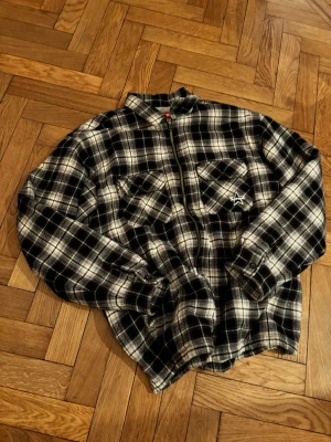 Svartvit rutig overshirt från Supreme - Supreme overshirt i svartvitt rutigt mönster med dragkedja framtill och två bröstfickor. Broderad Supreme-logga med stjärna på ena fickan. Overshirten är i mjukt, flanell-liknande material och har klassisk skjortkrage och knappar vid ärmslut.