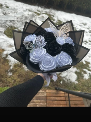 Bukett med svarta och vita rosor - Säljer en unik bukett med konstgjorda svarta och vita rosor, dekorerad med två dekorativa fjärilar i metall. Blommorna är inslagna i svart papper och buketten hålls ihop med ett vitt band. Perfekt som present eller dekoration.