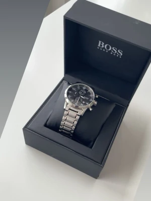 Hugo Boss silverfärgad klocka - Stilren klocka från Hugo Boss med silverfärgad länk i metall och svart urtavla. Klockan har kronograf-funktion, tydliga siffror och detaljer i vitt och silver. Perfekt accessoar för dig som gillar klassisk och modern stil. Och den tillverkas inte längre ! 
