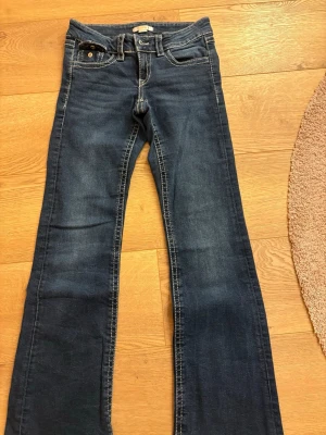 Mörkblå bootcut jeans med kontrastsömmar - Säljer ett par mörkblå bootcut jeans med vita kontrastsömmar och klassiska fem fickor. Jeansen har knapp- och dragkedjestängning samt ficklock med knapp bak. Perfekta för en avslappnad och trendig look.
