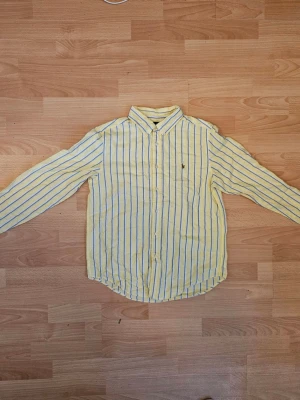 Gul och vit randig skjorta Ralph Lauren - Snygg gul skjorta från Ralph Lauren med vita och blå ränder. Klassisk krage, knäppning framtill och lång ärm. Liten broderad logga på bröstet. Perfekt för dig som vill ha en fräsch och stilren look.