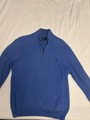Blå halvzip tröja från Polo Ralph Lauren - Snygg blå tröja från Polo Ralph Lauren med halv dragkedja och klassisk broderad logga på bröstet. Tillverkad i mjuk bomull och har ribbade muddar vid ärmslut och nederkant. Perfekt för en clean och stilren look. Väldigt bra skick.