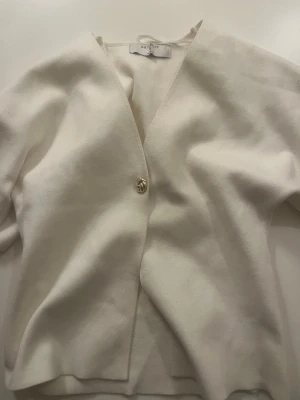 Vit kofta från Primark med knapp - Stilren vit kofta från Primark med en dekorativ guldfärgad knapp framtill. Koftan har V-ringning och är tillverkad i ett mjukt tyg som känns skönt mot huden. Perfekt för att lägga till en clean look till din outfit.