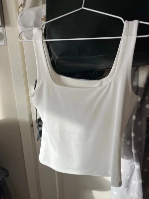 Vit ärmlös linnetopp - Snygg vit linnetopp med breda axelband och fyrkantig ringning. Toppen är enkel och stilren, perfekt att matcha med jeans eller kjol. Materialet känns mjukt och stretchigt, vilket ger en bekväm passform.