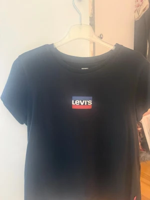 Svart Levi's t-shirt med logga - Snygg svart t-shirt från Levi's med klassisk logga i vitt, blått och rött på bröstet. T-shirten har rund halsringning och korta ärmar. Perfekt basplagg för en avslappnad och stilren look.