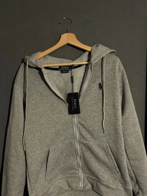Grå hoodie från Polo Ralph Lauren - Snygg grå hoodie från Polo Ralph Lauren med dragkedja framtill och klassisk huva med dragsko. Broderad logga i marinblått på bröstet och två fickor framtill. Tillverkad i mjuk bomullsblandning som känns skön mot huden. Perfekt för en avslappnad och stilren look.