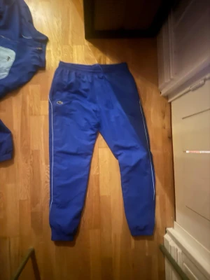 Blå träningsdress från Lacoste - Säljer en blå träningsdress från Lacoste med matchande byxor och jacka. Dressen har en cool tvåfärgad design i mörkblått och ljusblått, dragkedja på jackan och elastiska muddar. Perfekt för sport eller chill. Klassisk Lacoste-logga på både jacka och byxa.