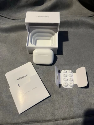 Apple AirPods Pro trådlösa hörlurar - Säljer ett par vita Apple AirPods Pro med laddningsetui, originalkartong, extra silikonpluggar och manual. Hörlurarna har en stilren och kompakt design i blank plast och levererar grymt ljud med brusreducering. Perfekta för musik, poddar och samtal.