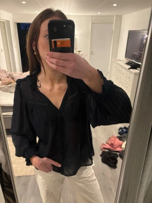 Svart transparent blus med volymärmar - Snygg svart blus i transparent material med v-ringning och knappar framtill. Blusen har långa, voluminösa ärmar med resår vid ärmslut och en lös passform. Säljer pga dålig användning tyvärr!❤️man kan även välja om man vill ha den knuten i halsen eller inte 