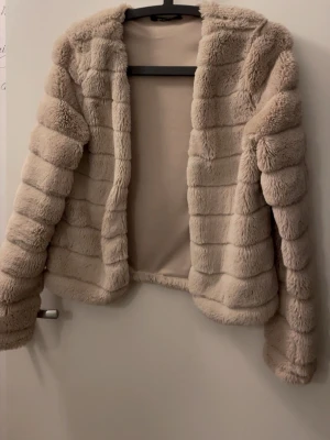 Beige fluffig pälsjacka - Säljer en supermjuk och fluffig beige pälsjacka med horisontella sömmar och öppen front. Jackan har lång ärm och en kortare, boxig passform som ger en trendig look. Perfekt för dig som vill ha en varm och stylish jacka till vintern.