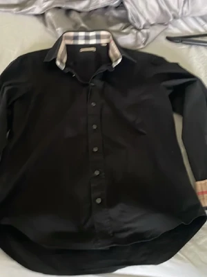 Svart skjorta från Burberry - Säljer en svart skjorta från Burberry med klassisk rutig detalj på insidan av kragen och manschetterna. Skjortan har knappar framtill och lång ärm. Perfekt för dig som gillar stilrena plagg med en twist.