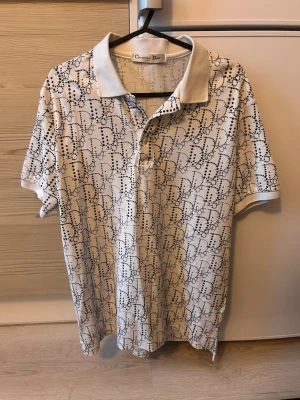 Vit mönstrad pikétröja  - Säljer en vit pikétröja  med svart prickigt mönster över hela plagget. Klassisk krage och knappar framtill. Perfekt för dig som gillar exklusiva märken och vill sticka ut med en unik topp.