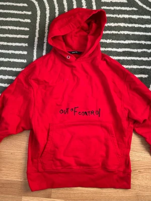 Röd hoodie från ZADIG&VOLTAIRE - Snygg röd hoodie från ZADIG&VOLTAIRE med svart broderad text 'OUT OF CONTROL' på bröstet. Klassisk känguruficka framtill och stor huva. Tillverkad i mjukt bomullsmaterial, perfekt för en avslappnad streetwear-look.