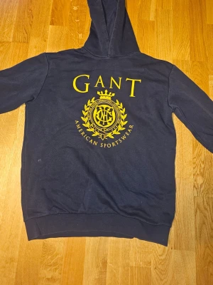 Marinblå hoodie från GANT - Snygg marinblå hoodie från GANT med stor gul logga. Strl 158-164. Riktigt bra skick!