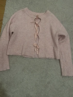 Rosa stickad kofta med rosetter - Söt ljusrosa stickad kofta med tre dekorativa rosetter framtill. Koftan har lång ärm, ribbstickad struktur och en något croppad passform. Perfekt för dig som gillar gulliga detaljer och mjuka material.