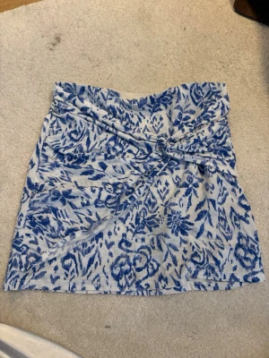 Blåvit blommig kortkjol med omlott - Kort kjol i blått och vitt med blommigt mönster och omlottdetalj framtill. Kjolen har en mjuk och lätt struktur, perfekt för varmare dagar. Ingen synlig logga eller märke.