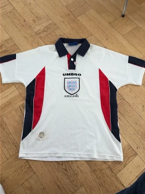ÄKTA vintage England fotbollströja från Umbro - Säljer en klassisk vit England fotbollströja från Umbro med blå och röda detaljer. Tröjan har korta ärmar, krage i mörkblått och tryckt England-emblem på bröstet. Perfekt för dig som älskar fotboll och vill ha en retro look.