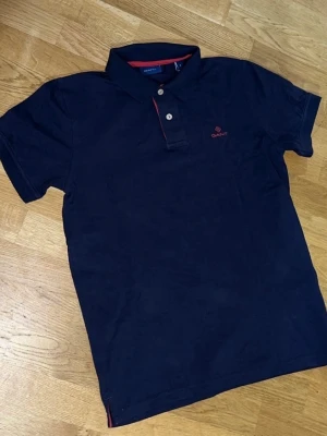 pikétröja från GANT - Klassisk mörkblå pikétröja från GANT med korta ärmar och krage. Tröjan har två knappar framtill och röd broderad logga på bröstet. Bra skick och passar till allt. Storlek: S