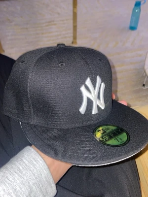 Svart New Era fitted Yankees keps 59FIFTY - Snygg svart keps från New Era 59FIFTY med klassisk New York Yankees-logga i vitt framtill. Platt skärm och broderade ventilationshål. Kepsen är i modellen fitted och har grå undersida på skärmen. Perfekt för dig som gillar streetwear och sportig stil.