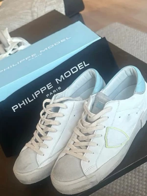 Vita Philippe Model sneakers med blå detalj - ANVÄNDA 2 GÅNGER HELT NYA  Snygga vita sneakers från Philippe Model med ljusblå detalj på hälen och grå mocka vid tån. Klassisk låg modell med vita skosnören och diskret logga på sidan. Perfekta för en stilren och trendig look.