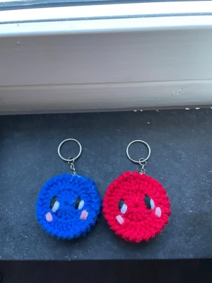Virkat nyckelring par smileys - Två virkade nyckelringar i form av runda smileys, en blå och en röd. Båda har broderade ögon och små rosa kinder. Perfekt att hänga på väskan eller nyckelknippan för en söt och personlig touch.