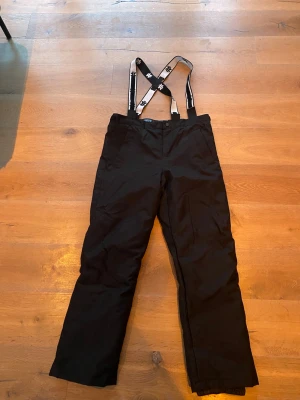 Svarta täckbyxor med hängslen - Svarta täckbyxor från Ski Industries med justerbara hängslen. Byxorna är vind- och vattenavvisande och har en vadderad insida för extra värme. Perfekta för kalla vinterdagar och skidåkning. Snygg och funktionell design med raka ben. Helt nya 