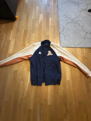 Blå och beige baseboll jacka Adidas - Snygg bomberjacka från Adidas i marinblått med beige och bruna ärmar. Jackan har broderad logga på bröstet och klassisk bomberkrage. Perfekt för dig som vill ha en sportig och trendig look.