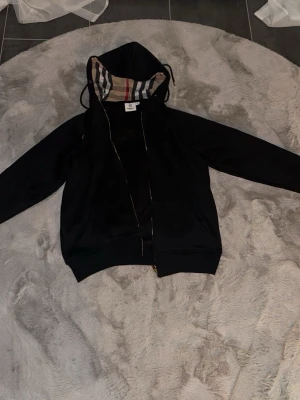 Svart hoodie från Burberry - Säljer en svart hoodie från Burberry med dragkedja och klassiskt rutigt foder i huvan. Hoodien har dragsko i huvan och diskret Burberry-logga broderad på bröstet. Perfekt för dig som gillar stilrena plagg med exklusiva detaljer. 