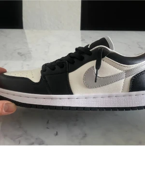 Nike Air Jordan 1 Low svart/vit - Snygga Nike Air Jordan 1 Low sneakers i svart och vitt med grå Swoosh. Klassisk siluett med lågt skaft, svart snörning och kontrasterande paneler. Ovandelen är i läder och sulan är vit med svart yttersula. Perfekta för dig som gillar streetwear och ikonisk design.