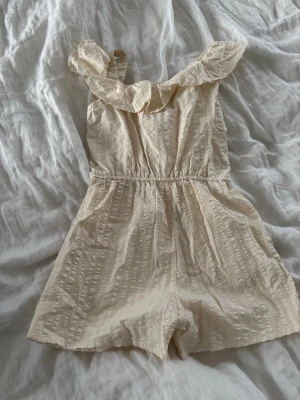 Beige off shoulder jumpsuit - Supersöt beige jumpsuit med off shoulder-krage och volangdetalj. Gjord i ett lätt, strukturerat tyg med resår i midjan för skön passform. Perfekt för sommarens alla äventyr och enkel att styla med sneakers eller sandaler.🤍🤍