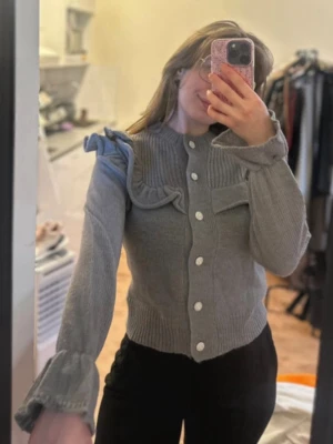 Grå stickad cardigan med volang - Grå stickad cardigan med vita knappar och volangdetaljer över axlarna. Tröjan har ribbstickad struktur upptill och långa ärmar med breda muddar. Perfekt för dig som gillar en trendig och feminin look.