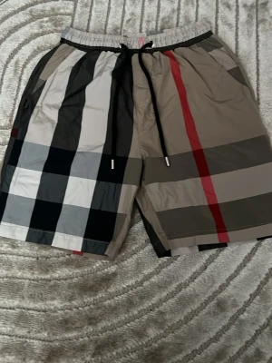 Burberry inspirerad shorts -  !Storlek M men den passar som S eller XS! Står inte Burberry på shorts. Snygga shorts med klassiskt rutigt mönster i svart, vitt, beige och rött. De har elastisk midja med svart snörning och praktiska fickor. Perfekta för sommaren och ger en trendig vibe till din outfit.