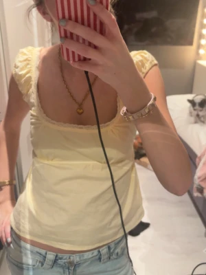 Gul blus med spets och rosett - Söt ljusgul blus med korta puffärmar, spetsdetaljer vid urringningen och en liten rosett i ryggen. Blusen har en feminin och luftig passform, perfekt till jeans eller kjol. Materialet känns som bomull och toppen har en romantisk vibe.