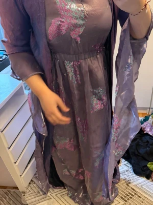 Lång kurdisk lila klänning med glittrigt mönster - Långklänning i lila med transparenta ärmar och glittriga mönster i rosa och blått. Klänningen har en lös passform och ett elegant fall, samt matchande sjal i samma material. Perfekt för dig som vill sticka ut med en unik look.
