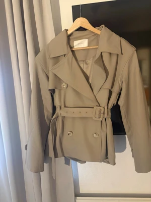 Beige trenchcoat från Meshki - Stilren beige trenchcoat från Meshki med klassisk dubbelknäppning, bred krage och bälte i midjan. Jackan har axelklaffar och dekorativa knappar, samt ett mjukt tyg som ger en snygg siluett. Perfekt för dig som gillar tidlös och trendig stil. Storlek xxs men är som s-m. Nypris 1900kr