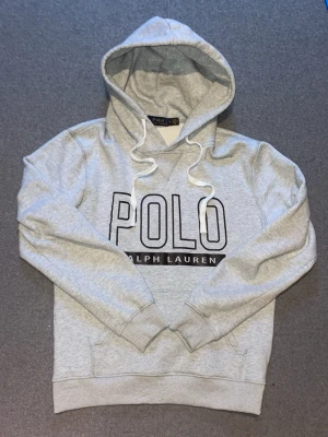 Grå hoodie från Polo Ralph Lauren - Snygg grå hoodie från Polo Ralph Lauren med stor logga framtill. Klassisk modell med huva och vita dragsnören samt magficka. Mjuk bomullsblandning och ribbade muddar vid ärmslut och nederkant. Perfekt för en avslappnad och trendig stil.