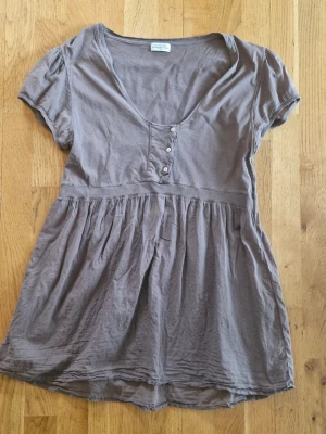 Hunkydory Sthlm Brun blus - Söt brun blus med korta puffärmar och v-ringning. Toppen har knappar framtill och ett rynkat parti under bysten som ger en avslappnad och flowy siluett. Perfekt till jeans eller kjol för en chill look.