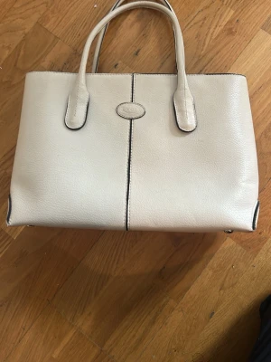 Beige handväska  - Stilren beige handväska med två handtag och diskret logga framtill. Väskan har en rektangulär form och svarta kontrastsömmar som ger en snygg detalj. Perfekt för dig som gillar klassisk och tidlös design.