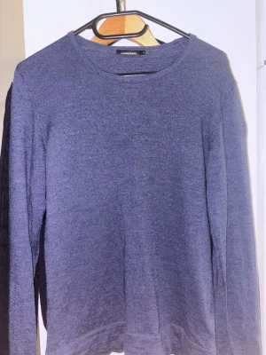 Mörkblå stickad tröja från J.Lindeberg - En stilren mörkblå stickad tröja från J.Lindeberg med rund halsringning och diskret logga på bröstet. Tröjan har långa ärmar och är tillverkad i mjukt stickat material som passar perfekt för lager-på-lager. Enkel och klassisk design.