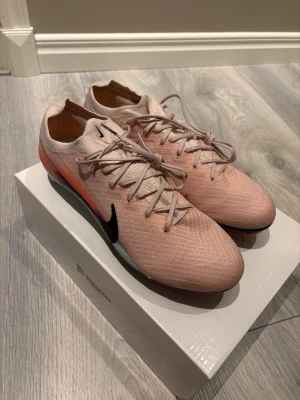 Nike Mercurial rosa fotbollsskor - Säljer ett par Nike Mercurial fotbollsskor i en snygg ljusrosa färg med svarta detaljer och orange inslag på sidorna. Skorna har en strömlinjeformad design, snörning och ovandel i textil för bra passform och känsla på planen. Perfekta för dig som vill sticka ut på matchen. 